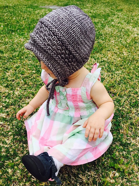Infant girl wearing knit Barre Bonnet and dress sitting on green grass.