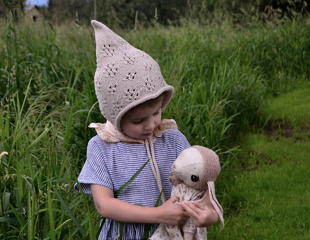 Young girl wearing hand-knit pixie bonnet and holding handmade bunny stuffed toy while standing in a field boarded by long grass.